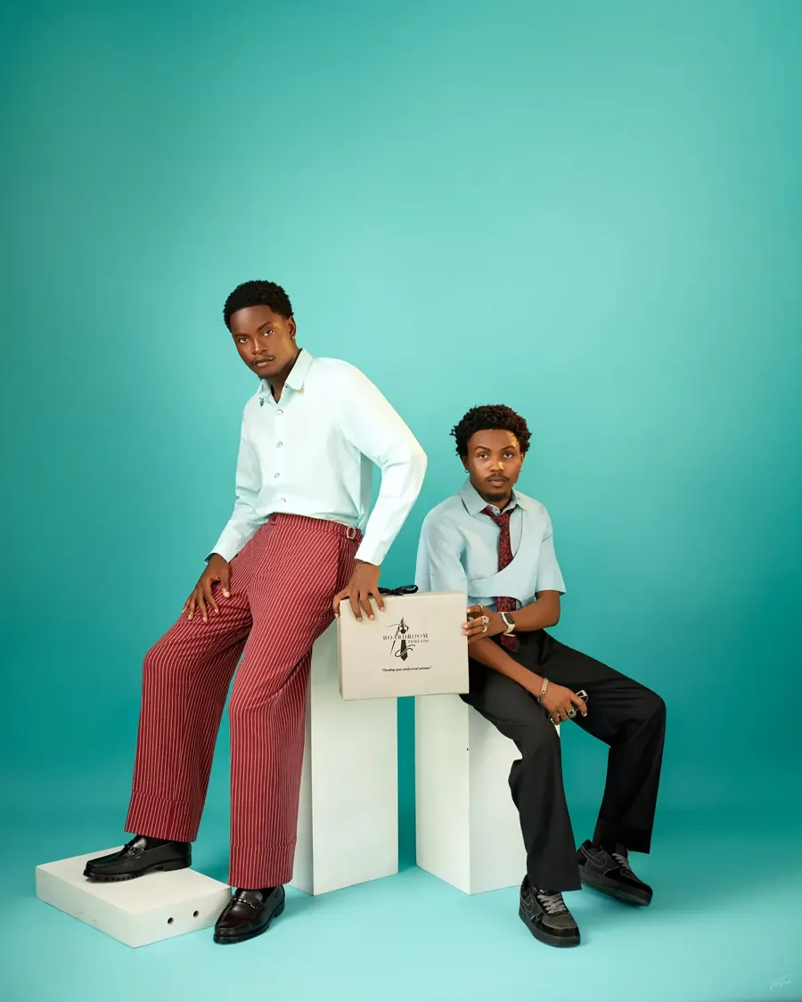Two models sitting on white plinths holding a Boardroom Threads box, one in burgundy pinstripe trousers and one in a short-sleeve shirt with a printed tie