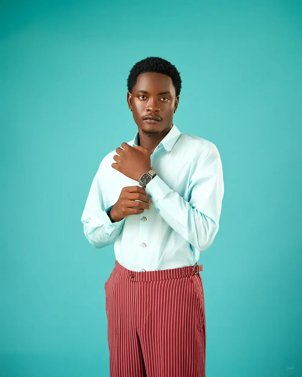 Model in a pale aqua shirt with eye-motif buttons and wide-leg burgundy pinstripe trousers, adjusting his watch against a teal backdrop