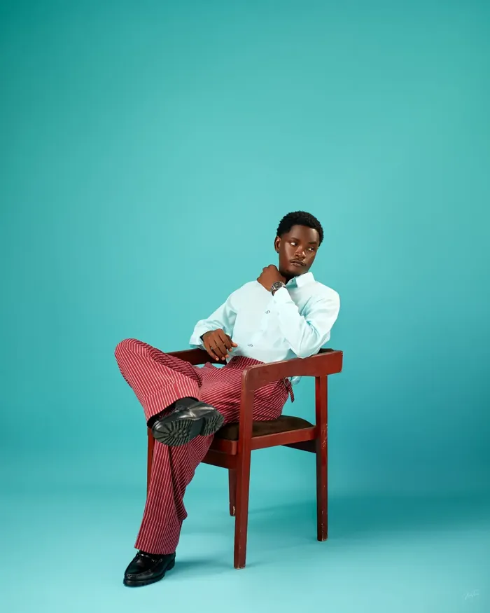 Model seated in a wooden armchair wearing burgundy pinstripe trousers and an aqua shirt